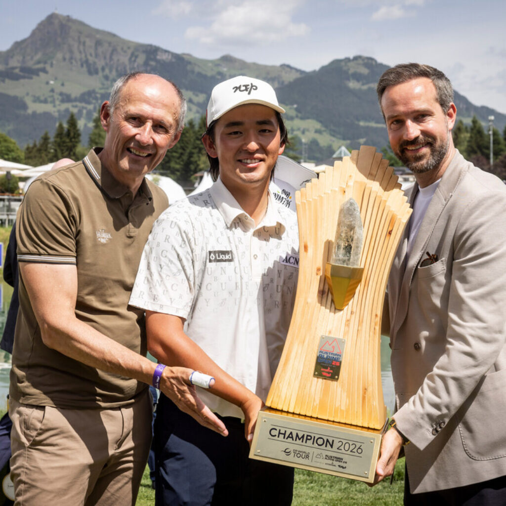 Drei Männer, die lächelnd eine große Trophäe in der Hand halten, mit Bergen und einem See im Hintergrund.