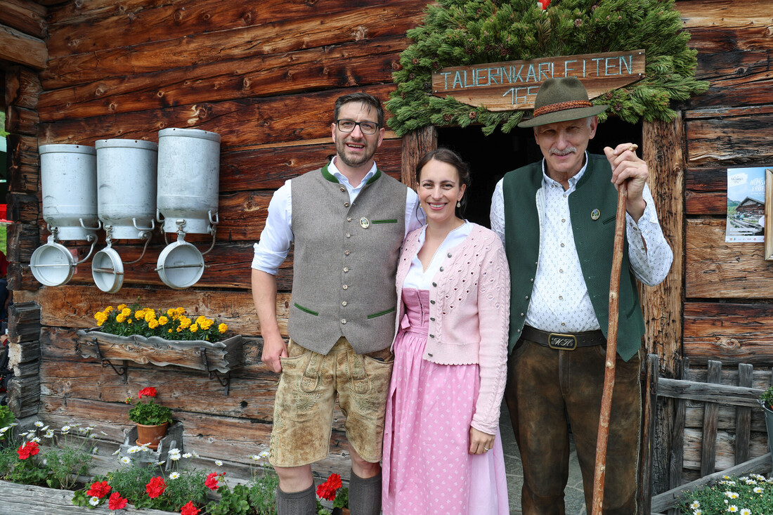 Drei Personen in traditioneller österreichischer Tracht stehen lächelnd vor einem rustikalen Holzhaus, das mit Blumen geschmückt ist.