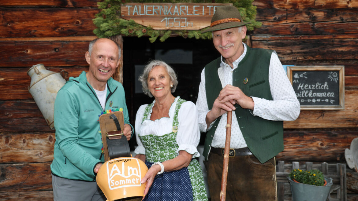 Drei lächelnde Menschen in traditioneller Alpenkleidung stehen vor einer rustikalen, geschmückten Holzhütte.