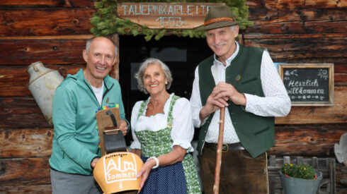 Drei lächelnde Menschen in traditioneller Alpenkleidung stehen vor einer rustikalen, geschmückten Holzhütte.