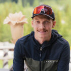 Ein Mann in Jacke und Mütze lächelt im Freien, im Hintergrund sind Bäume und eine Holzskulptur zu sehen.